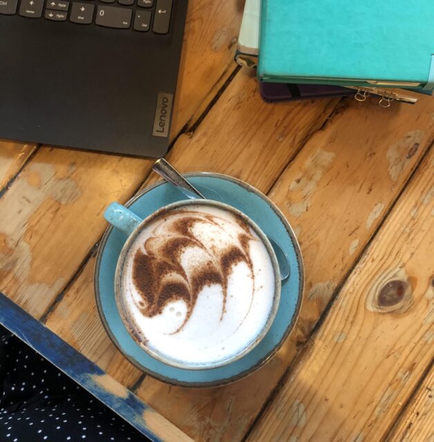 An overview shot of a funky chai latte in a turquoise cup and saucer. There's a wooded table top. There's a corner of a black laptop on the left and a stack of notebooks on the right. The notebooks are green, teal, and purple. Just imagine we're about to sit down to a discovery call together...
