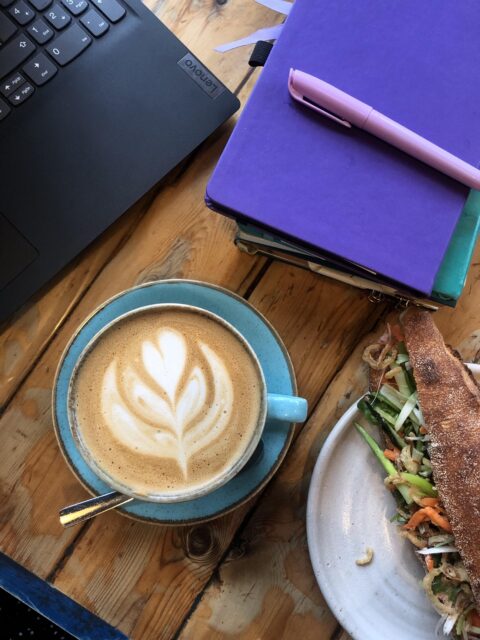 A close up with a coffee in a turquoise tea cup, a baguette with vibrant salad visible, a corner of a laptop, and a stack of notebooks with a purple hlighlighter on it.