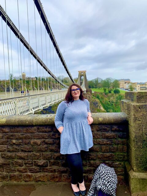 Bonnie the copywriter is stood in front of the Bristol Suspension Bridge. She's wearing a grey dress and black leggings. She is a white woman with long dark hair and chunky black glasses. 