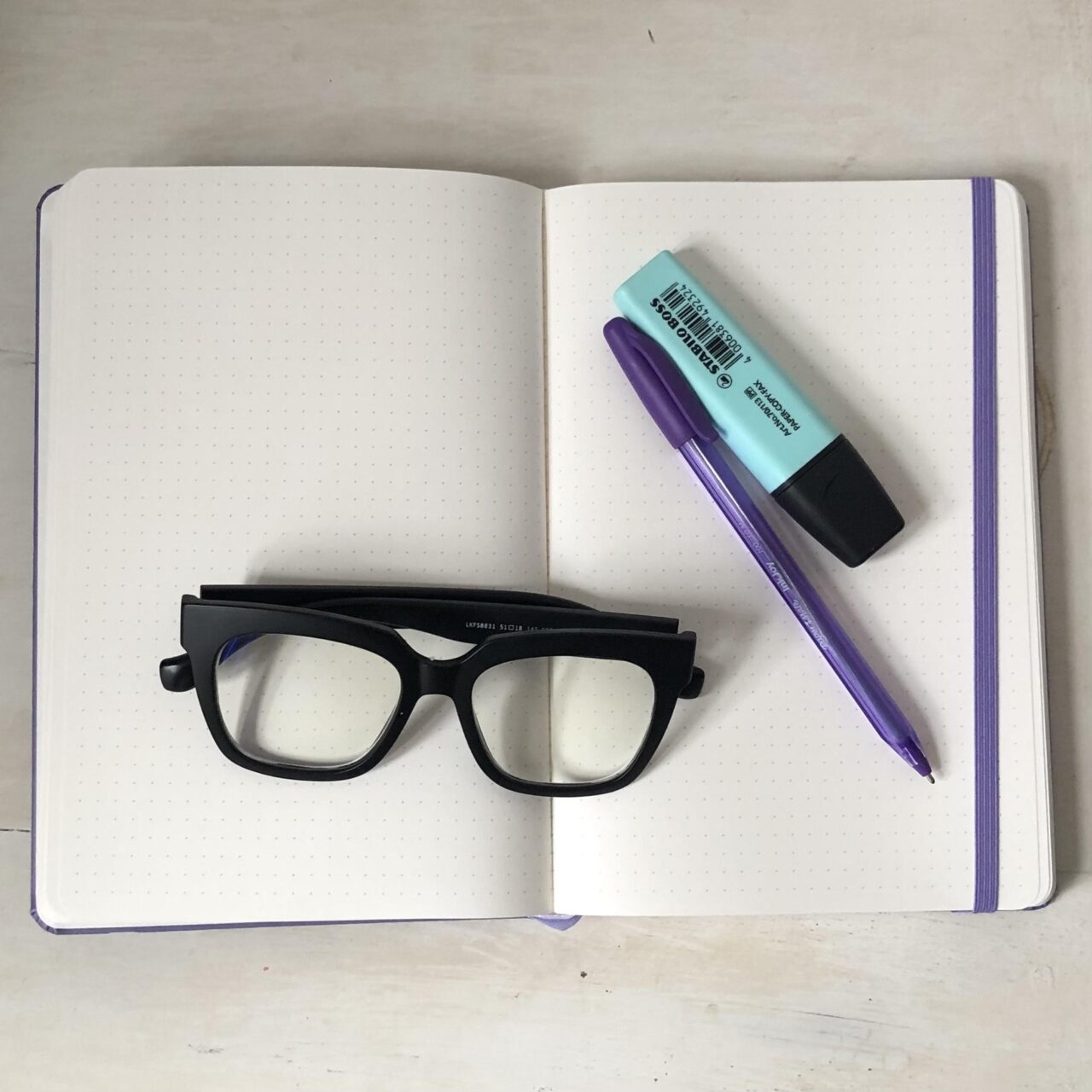 An open a5 notebook with Bonnie's black framed glasses on, and two pens, one purple biro and one green highlighter resting on the page.