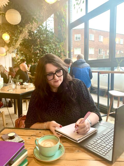 Copywriter Bonnie is writing in a notebook with her left hand. Theres a turquoise cup with coffee in in front of her, an open laptop, and a stack of notebooks. 