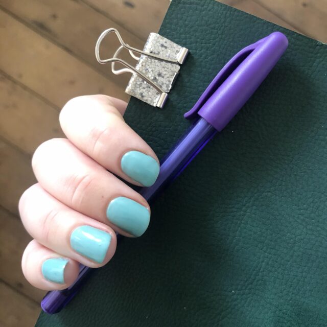 A close up of a white hand with turquoise painted fingernails. the hand is holding a purple pen, a green notebook, and there is a silver bulldog clip attached to the green notebook. 