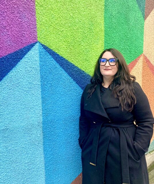 Bonnie, a white woman, stands in front of a super colourful graffiti wall