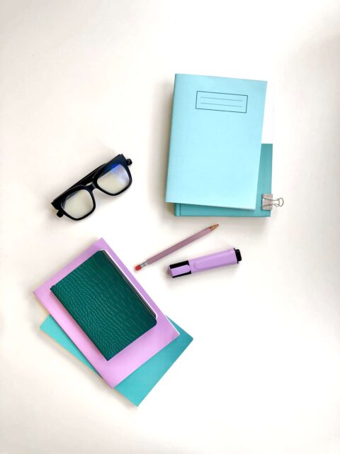 A flatlay of a bunch of stationery, there are some note books stacked up in colours of blue, purple, and green (the Words by Bonnie brand colours). There is also a pair of chunky black framed glasses and a purple highlighter. 