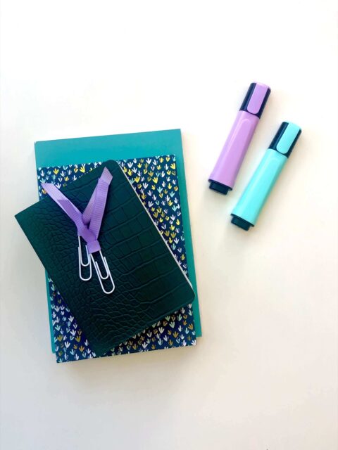 A white table top with three notebooks in colours of green and blue, a couple of paperclips with purple ribbons attached, and two highlighters, one green and purple. The perfect tools to plan for guest blogging and backlinks!