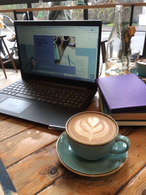 a table top with an open laptop showing a screen of website copy, a stack of notebooks (one purple one green) and a turquoise teacup and saucer with an oatmilk latte in it. Theres swirly latte art on the coffee.