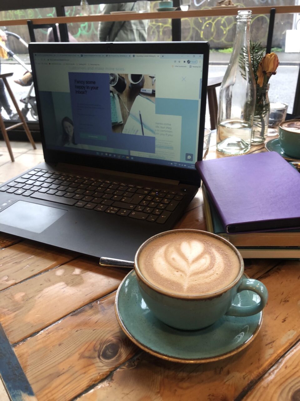a table top with an open laptop, a stack of notebooks (one purple one green) and a turquoise teacup and saucer with an oatmilk latte in it. Theres swirly latte art on the coffee.