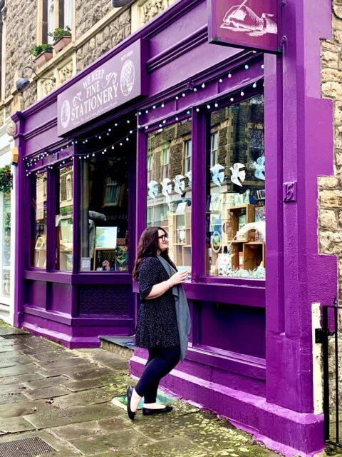 Copywriter Bonnie is standing in front of a stationery shop which is painted bright purple. Bonnie is a white woman with long dark hair. She's holding a turquoise travel coffee cup.
