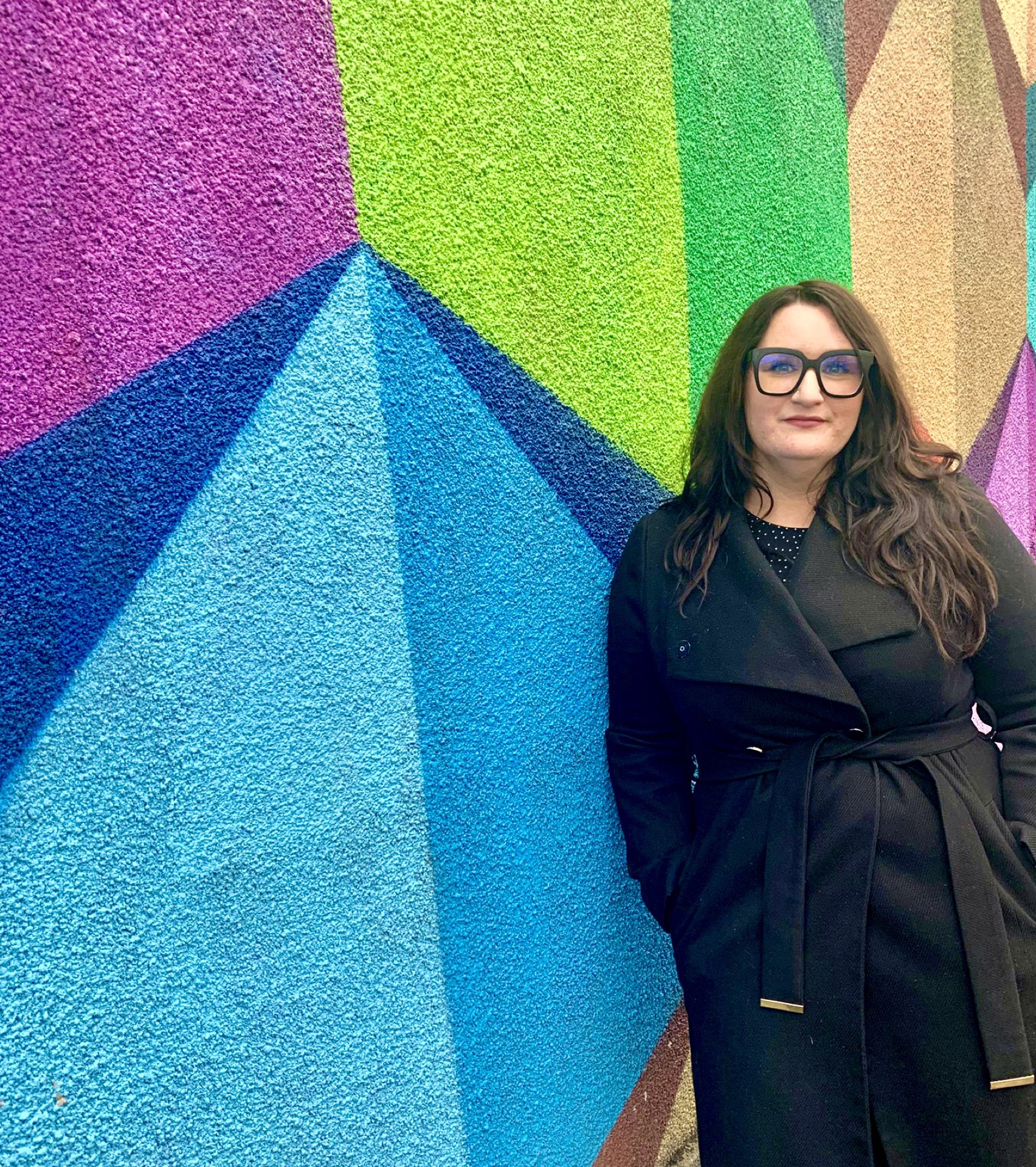 Copywriter Bonnie, a white woman with long dark hair, wearing a black coat, is leaning against a wall covered in graffiti. The graffiti is brightly coloured geometric shapes. 