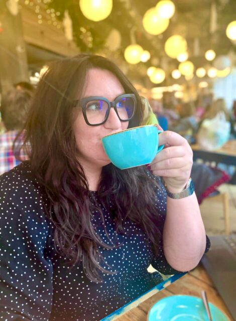 Bonnie, a white woman with long brown hair and black- framed glasses is looking away from the camera and drinking coffee from a turquoise cup.