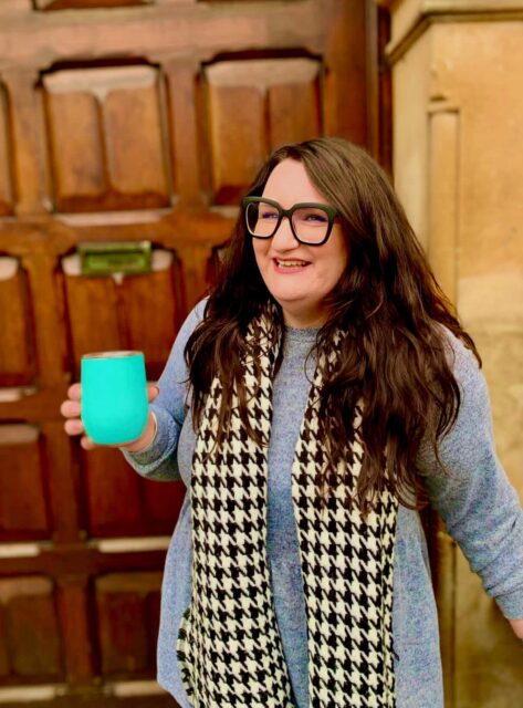 Bonnie, a white woman with long dark hair, is standing facing the camera, smiling, with a turquoise travel mug of coffee in her right hand. She's showing off that she's sustainable/ she cares about sustainability because she's got a travel mug. 