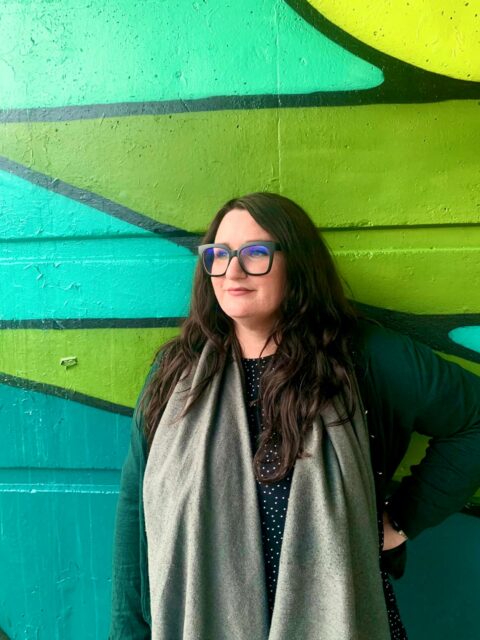 Bonnie- a white woman with long dark hair and chunky glasses- is stood in front of a spray painted wall looking towards her right, perhaps all all the lovely copywriting clients she has as a result of this blog. 