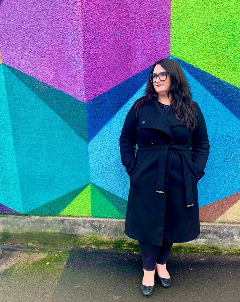 Copywriter Bonnie is standing in front of a wall of graffiti in Bristol. The wall is covered in large geometric shapes in purple, teal, green, turquoise and dark blue. Bonnie is a white woman with long dark hair, wearing a black coat. Used for the "small business blog" blog.