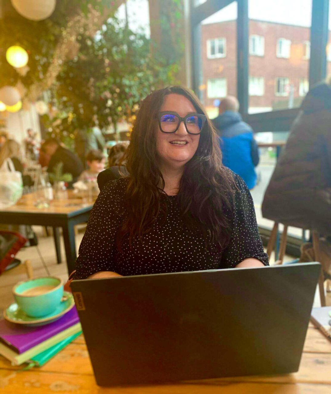 Copywriter Bonnie is sat in front of a laptop smiling. Bonnie is a white woman with long dark hair and chunky black framed glasses. She's probably writing an SEO blog for a copywriting client.
