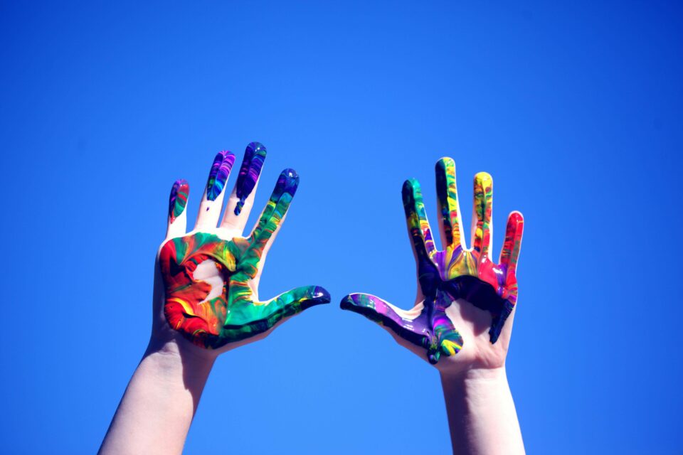 Against a bright blue sky two hands are raised up. They belong to a white person, perhaps a child, and there are smears of paint making a rainbow on the palms. Image used here to represent LGBTQ+ families.