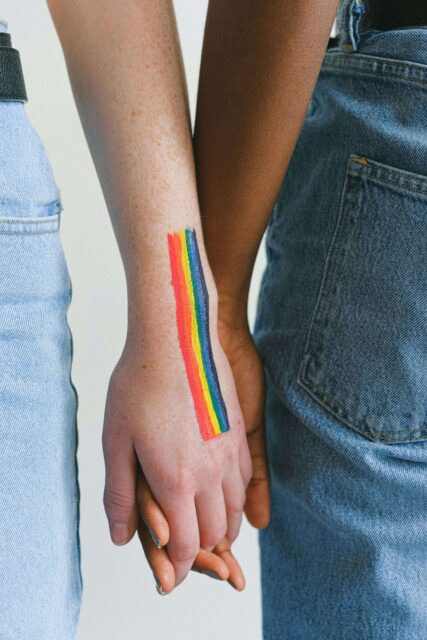A close up of two hands. Two people are holding hands and one hand has a long rainbow painted on it. This image is being used to represent LGBTQ+ relationships.