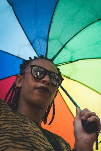 An image looking up at a female- presenting person wearing sunglasses and looking cool AF. She's holding a rainbow umbrella open behind her. 