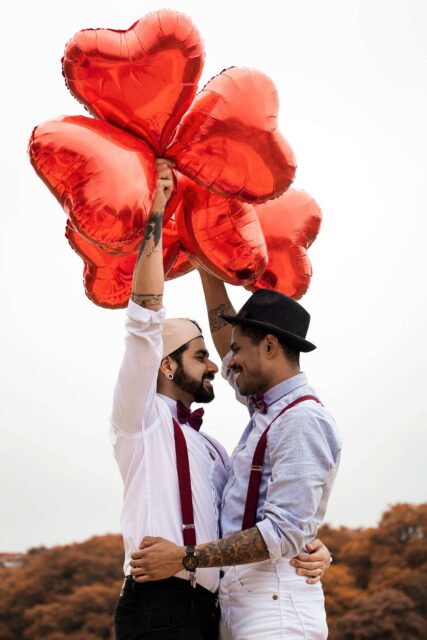 A happy couple on their wedding day. Two male- presenting people are staring into each other's eyes, with their hands held above them holding red, helium, love- heart balloons. This image is representing LGBTQ+ weddings. 