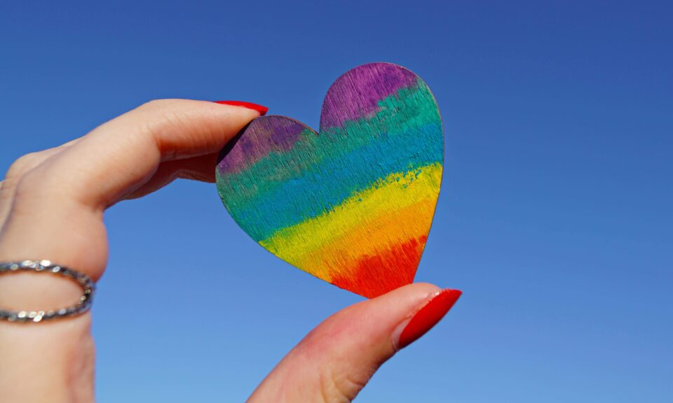 A close up of a thumb and finger holding a wooden heart which has been coloured with chalk in rainbow colours. This image is being used to represent LGBTQ+ related products. 