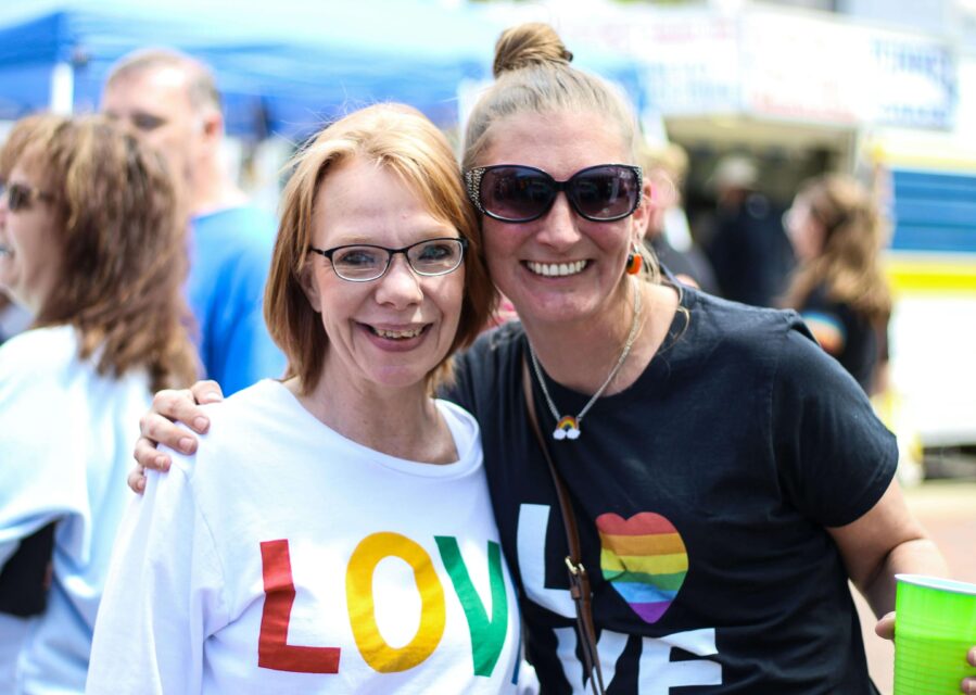 A close up of two female- presenting people, embracing, facing the camera, and smiling. They wear t-shirts with 'Love is love' in rainbow colours. 