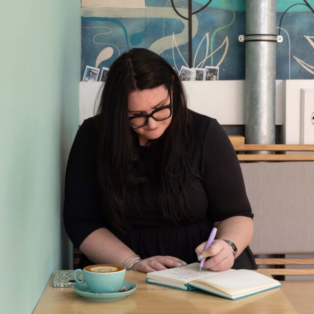 Bonnie, a white woman with long brown hair and chunky framed glasses, is sat in a cafe hand writing in a notebook. 