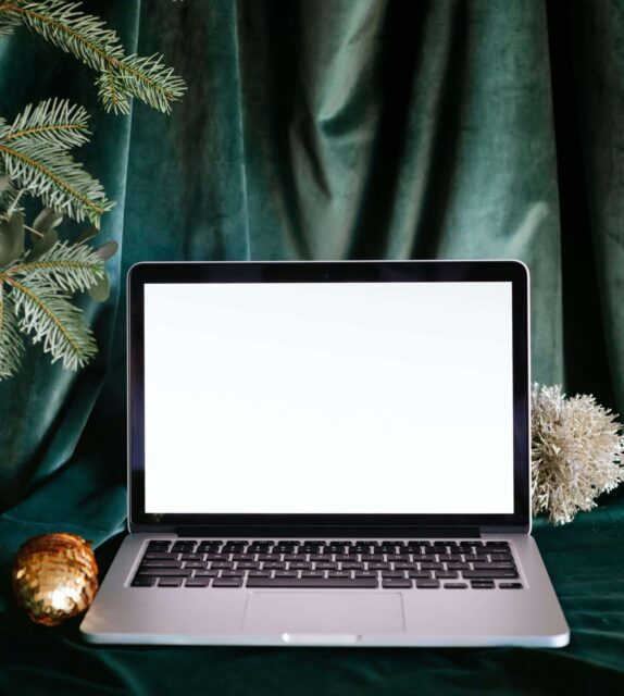 An open laptop in front of a green velvet fabric backdrop and some Christmas tree branches. It's not very "Christmas in july" tbh but it does have "christmas/ writing" vibes.