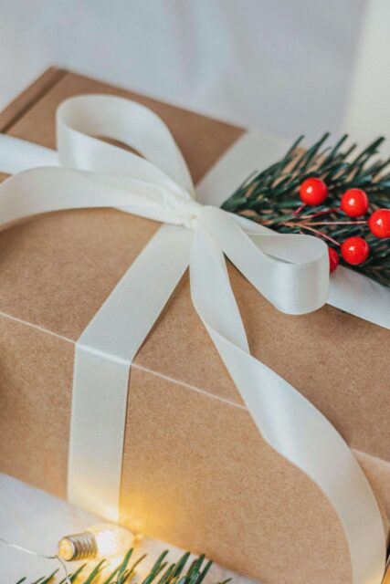 Close up of a gift wrapped in brown paper with a white satin ribbon. Tucked into the ribbon is a green branch with red berries. Subtle Christmas vibes. 