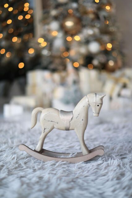 In the centre of the image is a little mini rocking horse. In the distance all blurry are some Christmas gifts and some tree branches and fairy lights